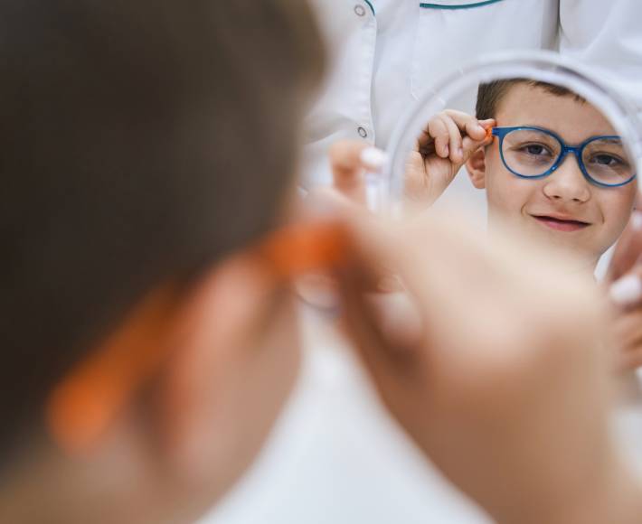 Cute male child trying on eyeglasses and smiling while having appointment with doctor optometrist